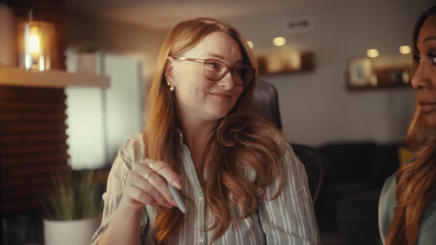 A woman with glasses smiles and listens intently while collaborating with a colleague in a modern home office