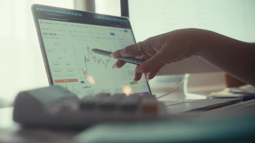A financial analyst points with a pen to a fluctuating stock market chart on a laptop screen while working from a home office