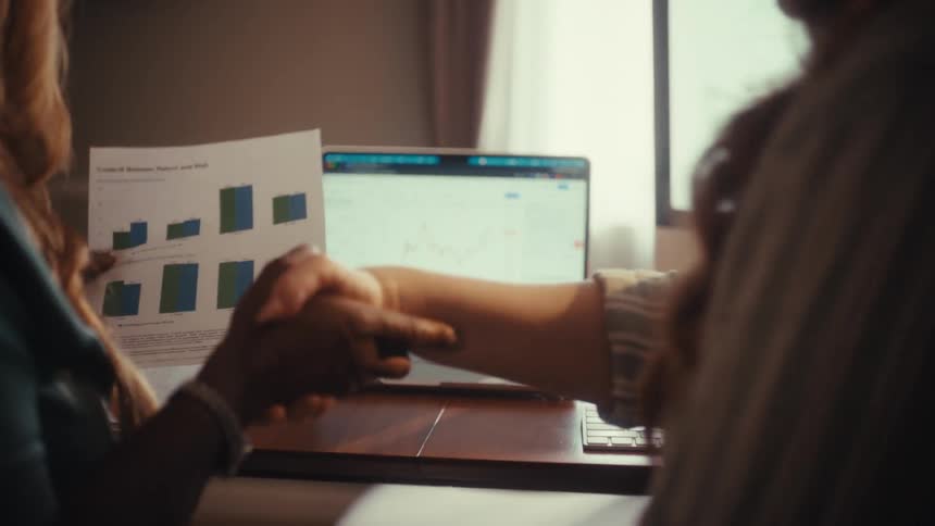 Over-the-shoulder shot of two businesswomen analyzing financial charts and data, shaking hands to close a successful deal