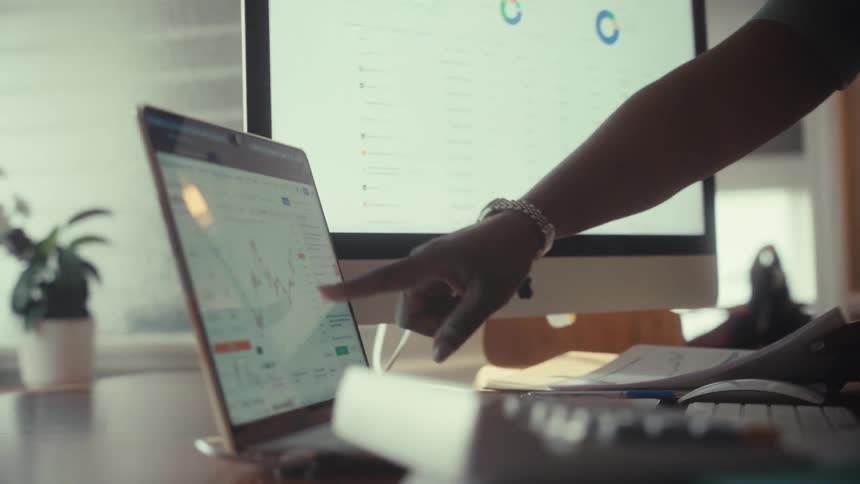 Two female colleagues collaborate while analyzing financial charts and stock market data on their computers in a modern office