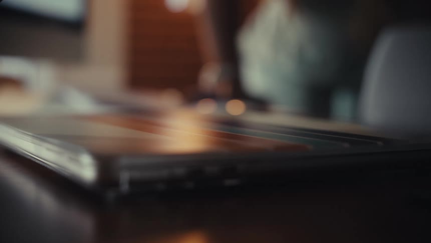 A soft focus, close-up shot of a person's hand opening a laptop on a wooden desk to begin working