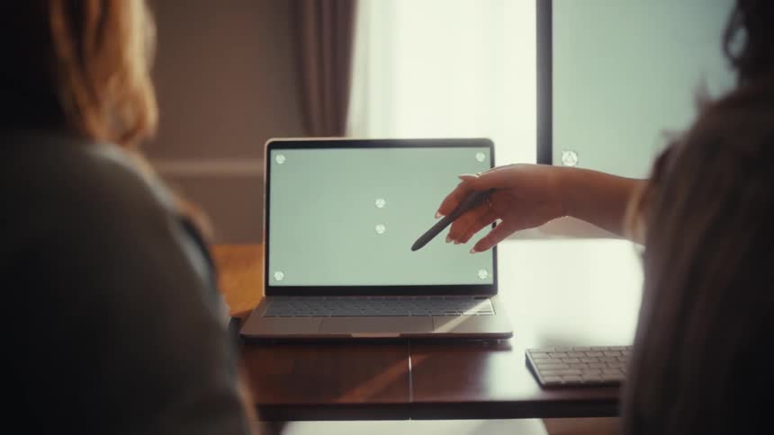 Two women collaborate in a sunlit office, with one pointing a stylus at a laptop's green screen with tracking markers