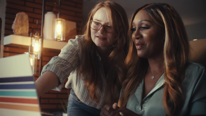 Two diverse female colleagues collaborate on a project, pointing at a laptop screen in a warmly lit office