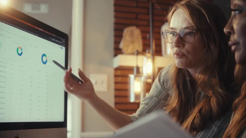 A young businesswoman wearing glasses explains financial data and charts on a computer screen to her colleague in a collaborative office meeting