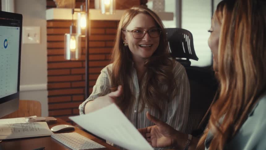 Two diverse businesswomen collaborate in an office, reviewing data on a computer before smiling and shaking hands to finalize a successful deal