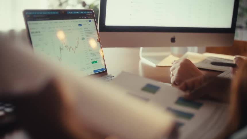A financial analyst reviews printed charts while monitoring the stock market on a laptop at their desk