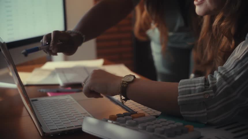 Two diverse businesswomen collaborate in an office, pointing at charts and data on their computers in slow motion