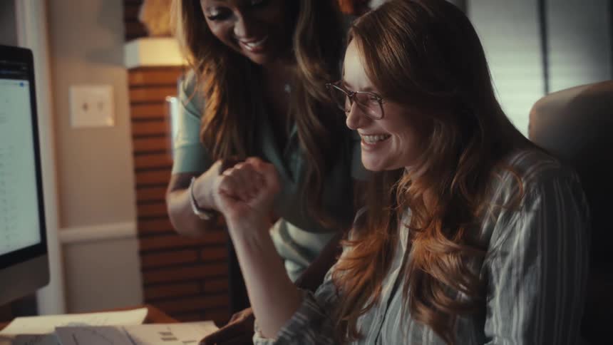 Two diverse businesswomen celebrate a professional success while working together at a computer in a cozy home office