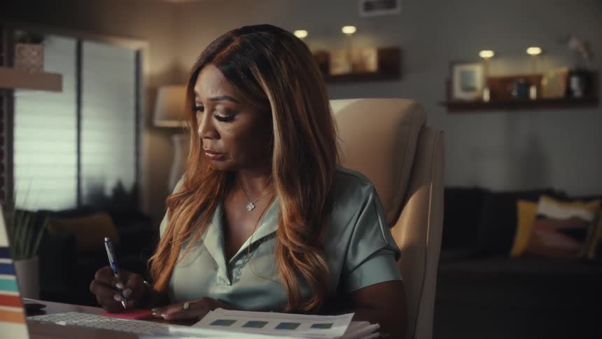 A focused Black woman works diligently from her home office, taking notes while looking at her laptop