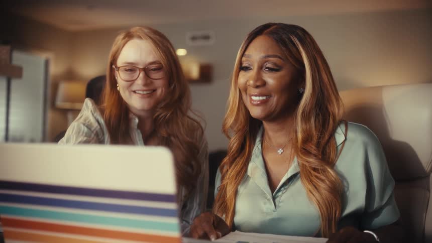 Two happy and diverse female colleagues laugh while working together on a laptop in a warmly lit home office