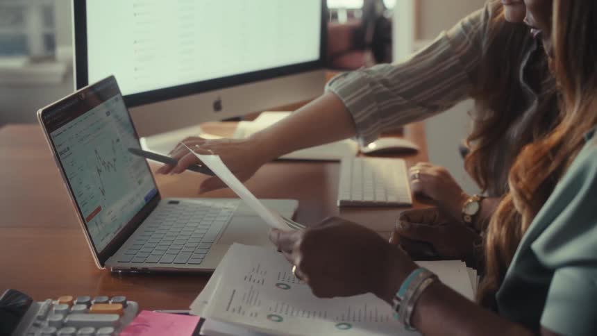 Two female colleagues collaborate at a desk, comparing financial charts on paper with a stock market graph on a laptop