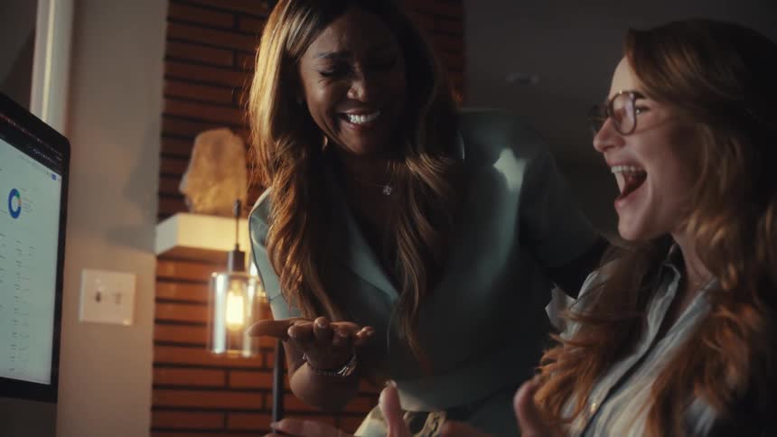 Two diverse female colleagues joyfully celebrate a business success while analyzing charts and data on a computer screen in their office