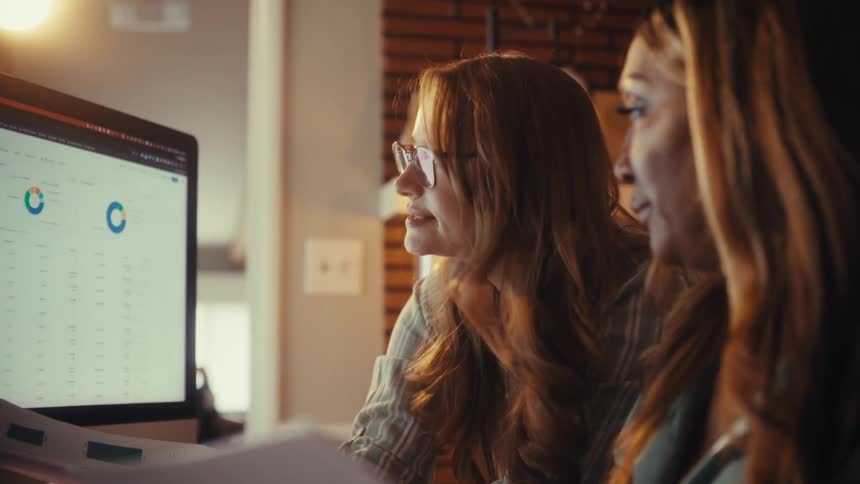 Two diverse female colleagues collaborate, analyzing business charts and data together on a computer screen in a cozy, modern office