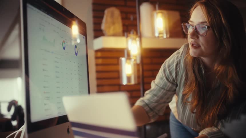 Two diverse women collaborate on a project, pointing at graphs and data on a computer screen in a cozy, modern office