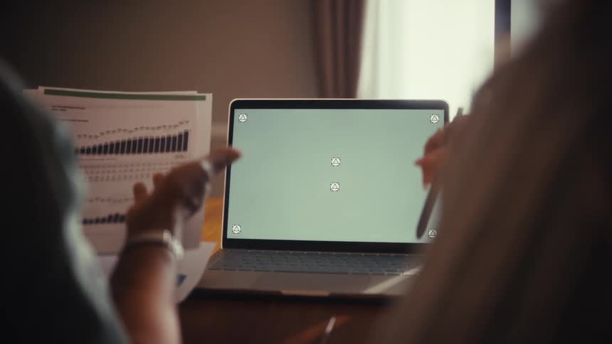 Over the shoulder view of two businesswomen collaborating over a laptop with a green screen, analyzing financial charts and data
