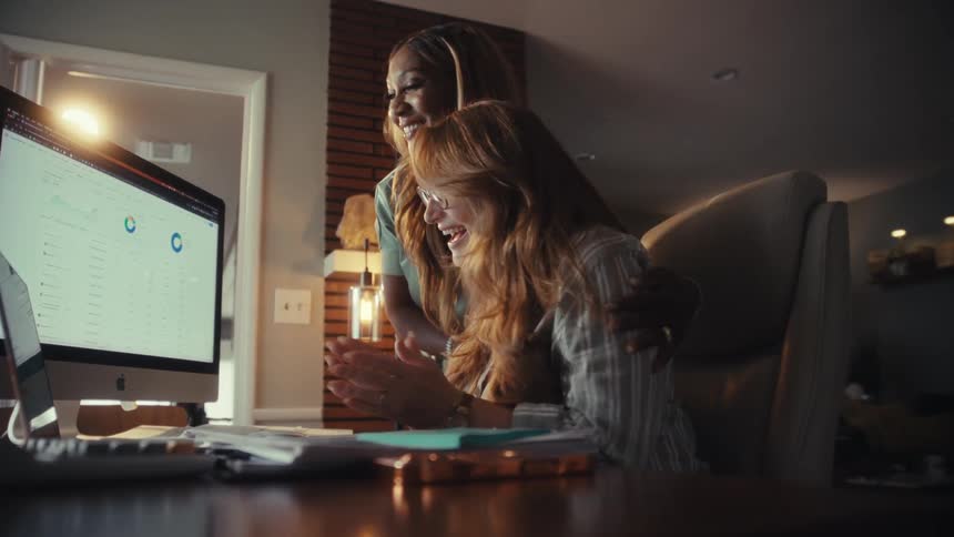 Two diverse female entrepreneurs smile and celebrate a successful outcome while analyzing data charts on a computer in their home office