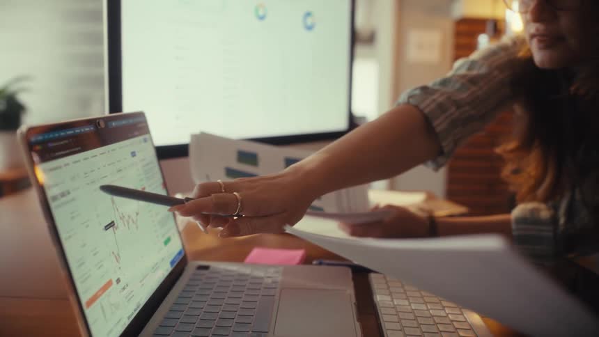 A financial analyst points at a stock market chart on her laptop while comparing it with paper documents in her home office