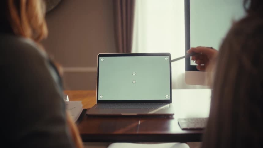 Over the shoulder view of two female colleagues collaborating on a project using a laptop and desktop computer with green screens in a home office