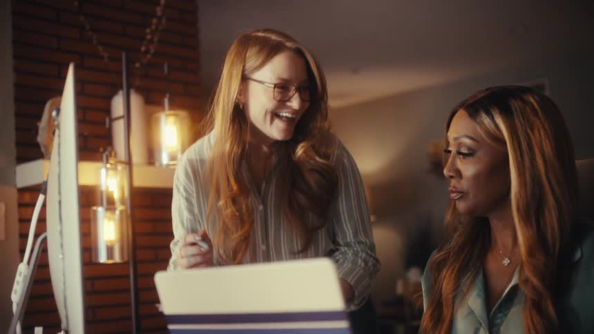 Two diverse female entrepreneurs smile while collaborating on a project in a modern home office at night