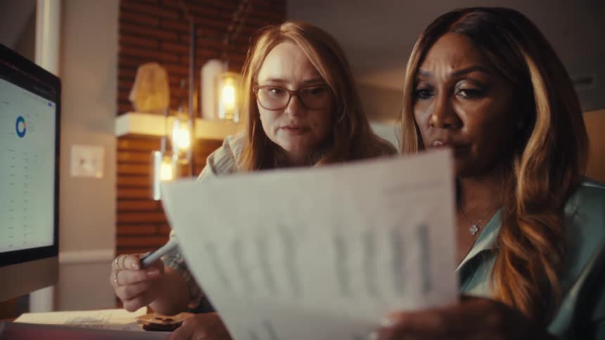 Two focused businesswomen collaborate in a modern office, analyzing financial charts on a computer screen and a printed report