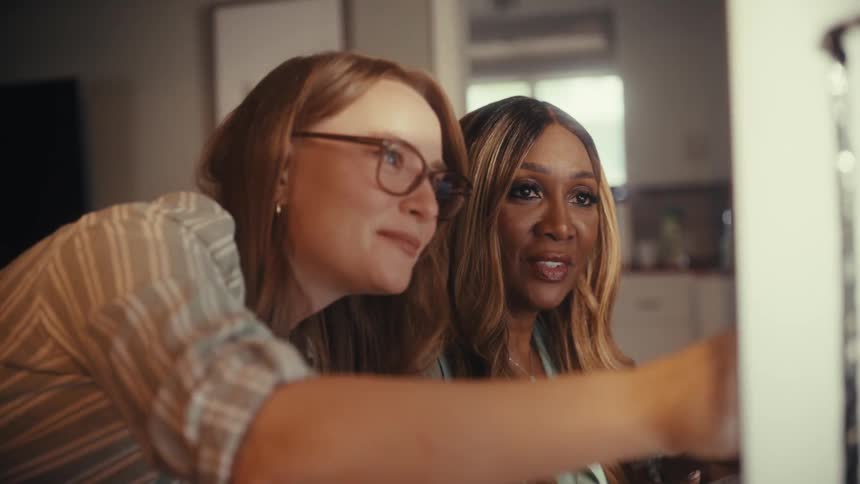 Two diverse female colleagues smile as they collaborate on a project, looking intently at a computer screen in their office