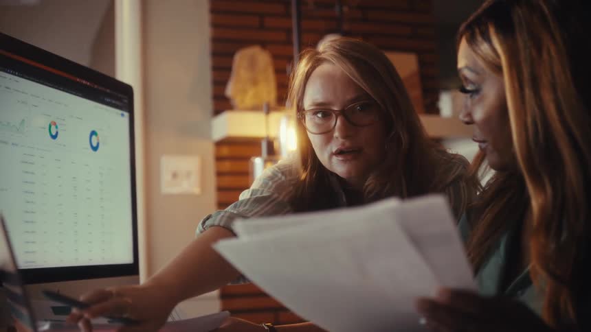 Two diverse female colleagues collaborate in a modern office, analyzing financial data and charts on a computer screen