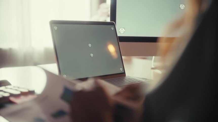 An over-the-shoulder shot shows a professional pointing at a green screen laptop while reviewing financial charts at their desk