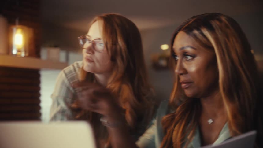 Two diverse female coworkers collaboratively discuss a project, pointing at a computer screen in a warmly lit modern office