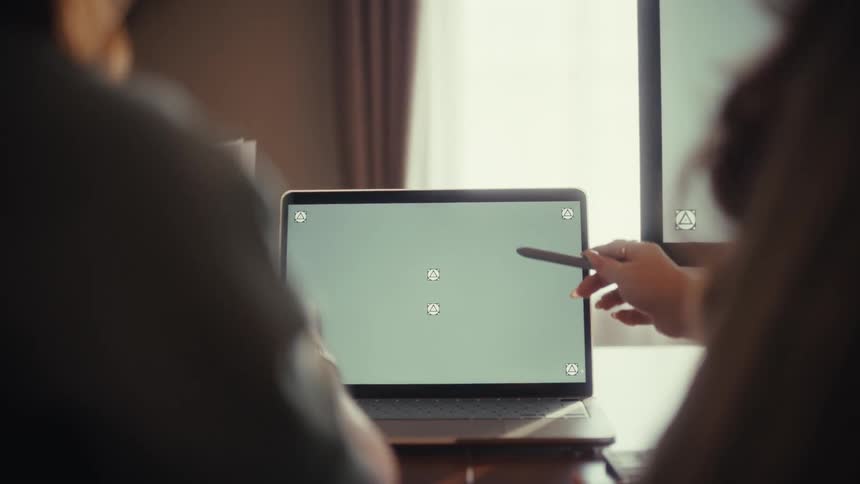 Over-the-shoulder shot of two colleagues collaborating and pointing at a laptop with a green screen in a modern office