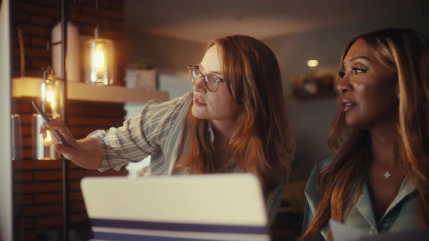 Two diverse female colleagues collaborate on a project, analyzing data on a computer screen late at night in a cozy home office