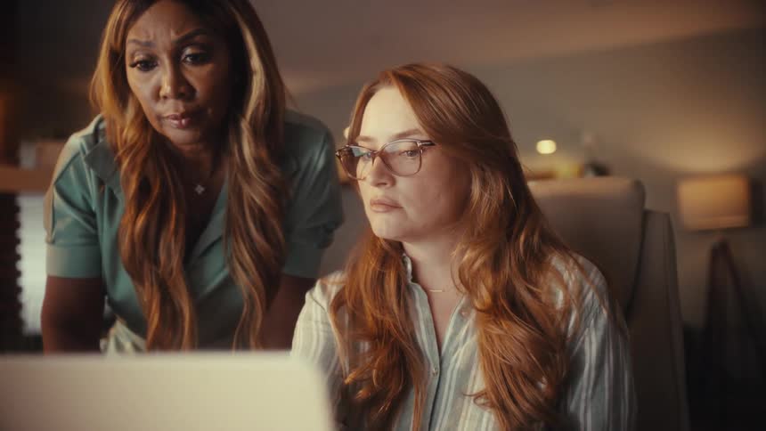Two diverse female colleagues collaborate on a project, intensely focused on a laptop screen in a warmly lit office at night