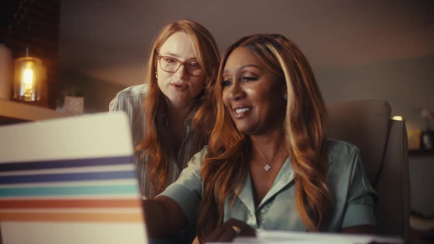 Two diverse and happy women collaborate on a laptop in a cozy home office at night