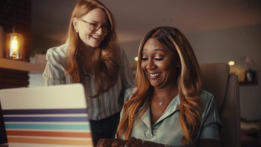 Two diverse female colleagues smile as they collaborate on a project using a laptop in a warm, modern office at night
