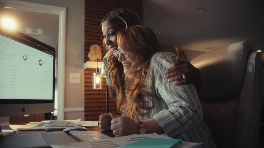 Two diverse female colleagues joyfully celebrate a business success while analyzing data on a computer in a home office