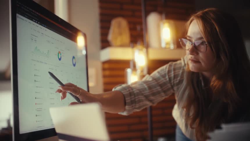 A focused businesswoman with glasses points to a data dashboard on a computer screen, collaborating with her colleague in a modern office