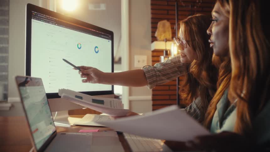 Two female colleagues collaborate while analyzing data and graphs on a computer in their cozy home office