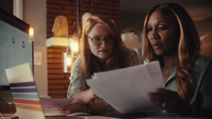 Two diverse female colleagues work together in a home office, discussing business analytics on a computer while reviewing documents