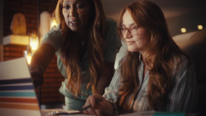 Two diverse businesswomen collaborate on a project, pointing at a laptop screen while working late in a warmly lit office