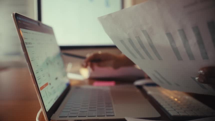 Two businesswomen analyze financial charts, comparing a printed report with live data on a laptop in their office