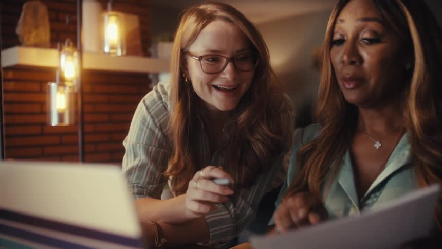 Two diverse female colleagues collaborate on a project late at night, reviewing data on a laptop and paper documents in a cozy home office