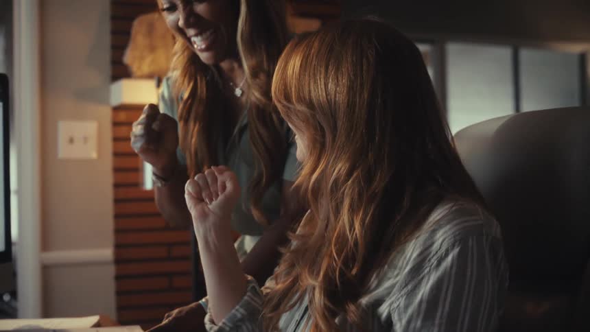 Two diverse businesswomen joyfully celebrate a project's success while collaborating on a computer in a modern office at night