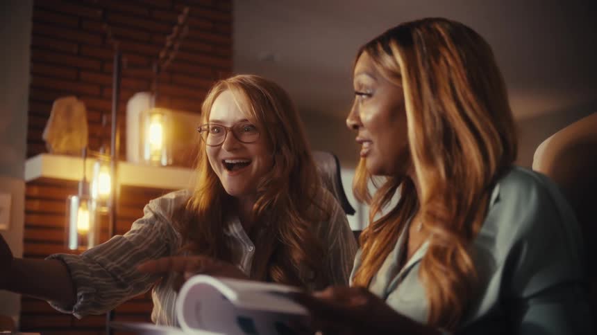 Two diverse female coworkers review project documents together in a modern office, celebrating their successful collaboration with a joyful handshake