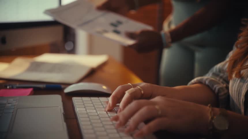 A slow motion close-up shows a woman's hands typing on a keyboard while a colleague in the background reviews business reports