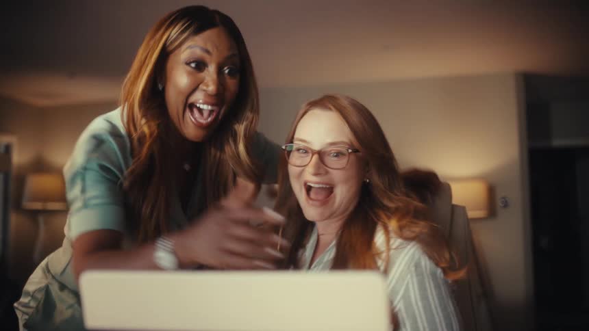 Two diverse and excited businesswomen celebrate a shared success on their laptop at night, culminating in a joyful hug and laughter