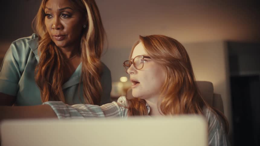 Two diverse businesswomen work together late at night, focused on a computer screen in a home office