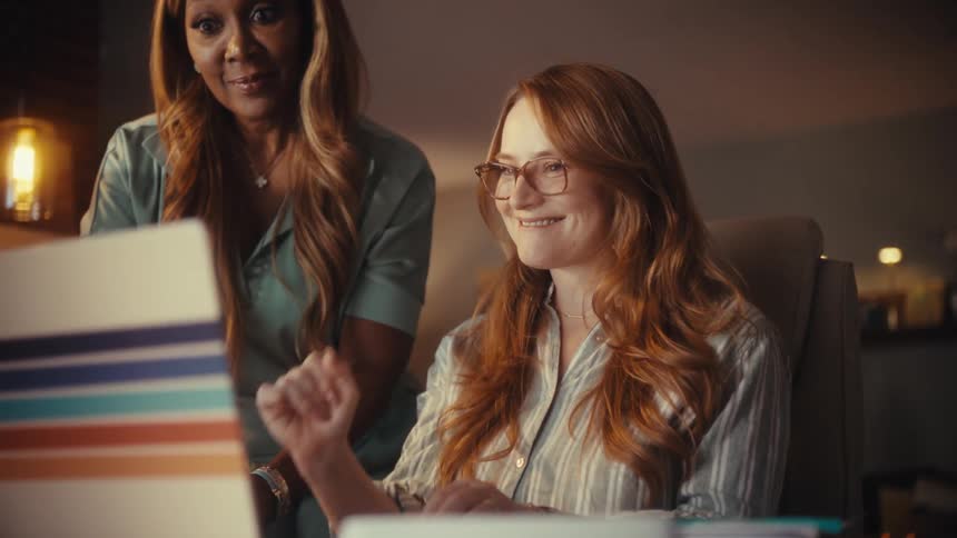 Two diverse professional women working late at night in their home office celebrate a successful project with smiles and a fist pump