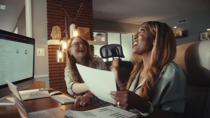 Two diverse businesswomen laugh and shake hands in celebration of a successful deal while working together in a modern home office