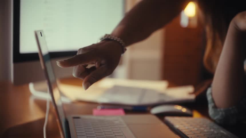 A close-up in slow motion of two diverse female colleagues analyzing data charts on a laptop and desktop computer in a warm-lit office