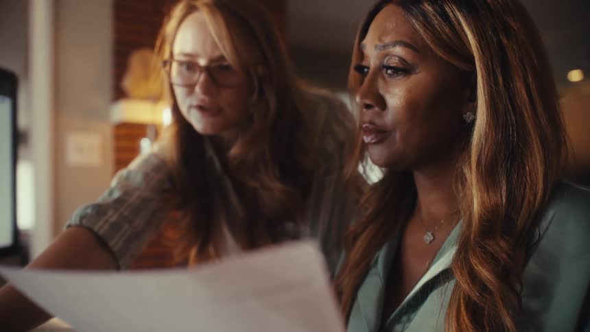 A slow rack focus reveals two diverse professional women collaborating and reviewing financial documents together in a home office