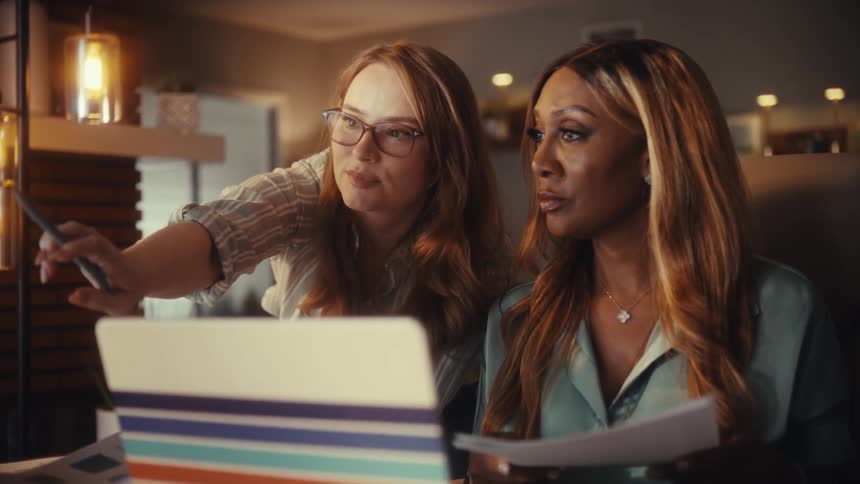 Two diverse female colleagues collaborate on a project in a warmly lit home office, pointing at a computer screen and discussing details